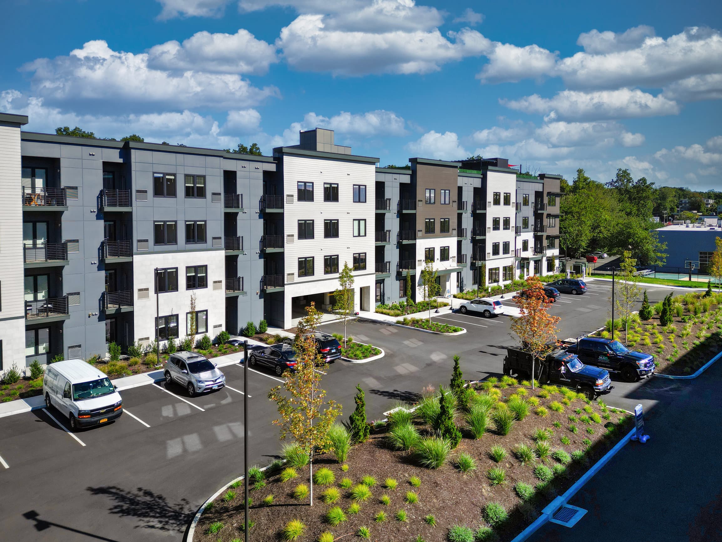 Modern apartment buildings with balconies, a parking lot, and landscaped shrubs under a blue sky with clouds