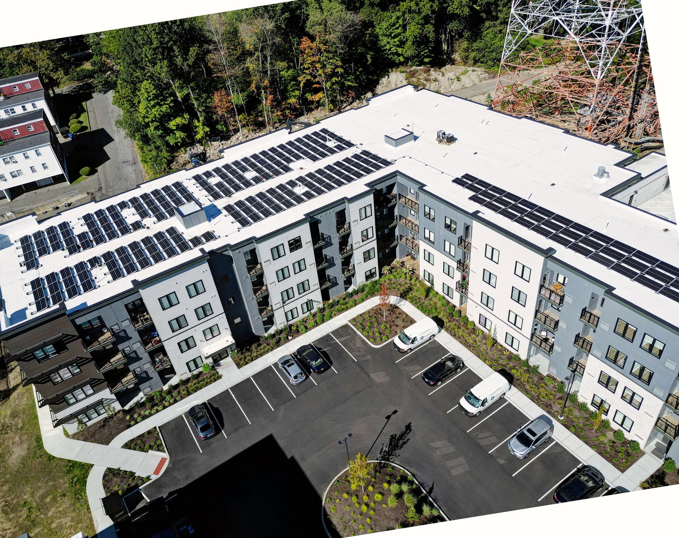 Aerial view of a white apartment building with solar panels on the roof, a parking lot and a lattice tower