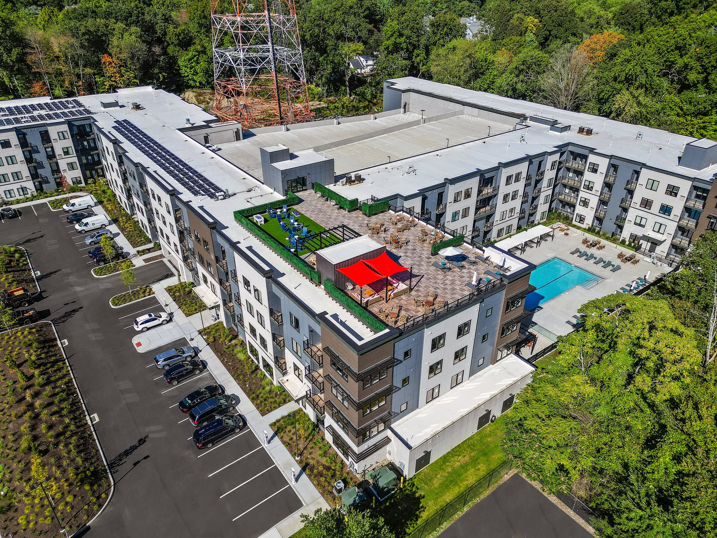 Aerial view of apartment complex with solar panels, terrace seating, a pool, a red lattice tower, and trees