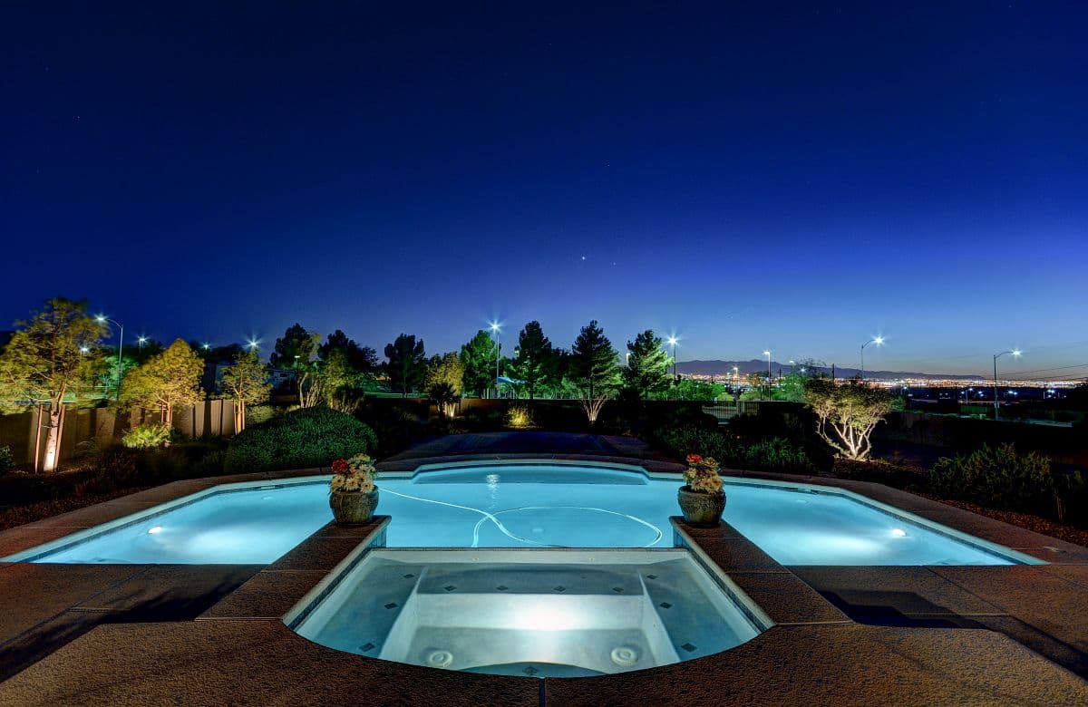 Night view of an illuminated pool and spa with two planter pots, surrounding trees and distant city lights