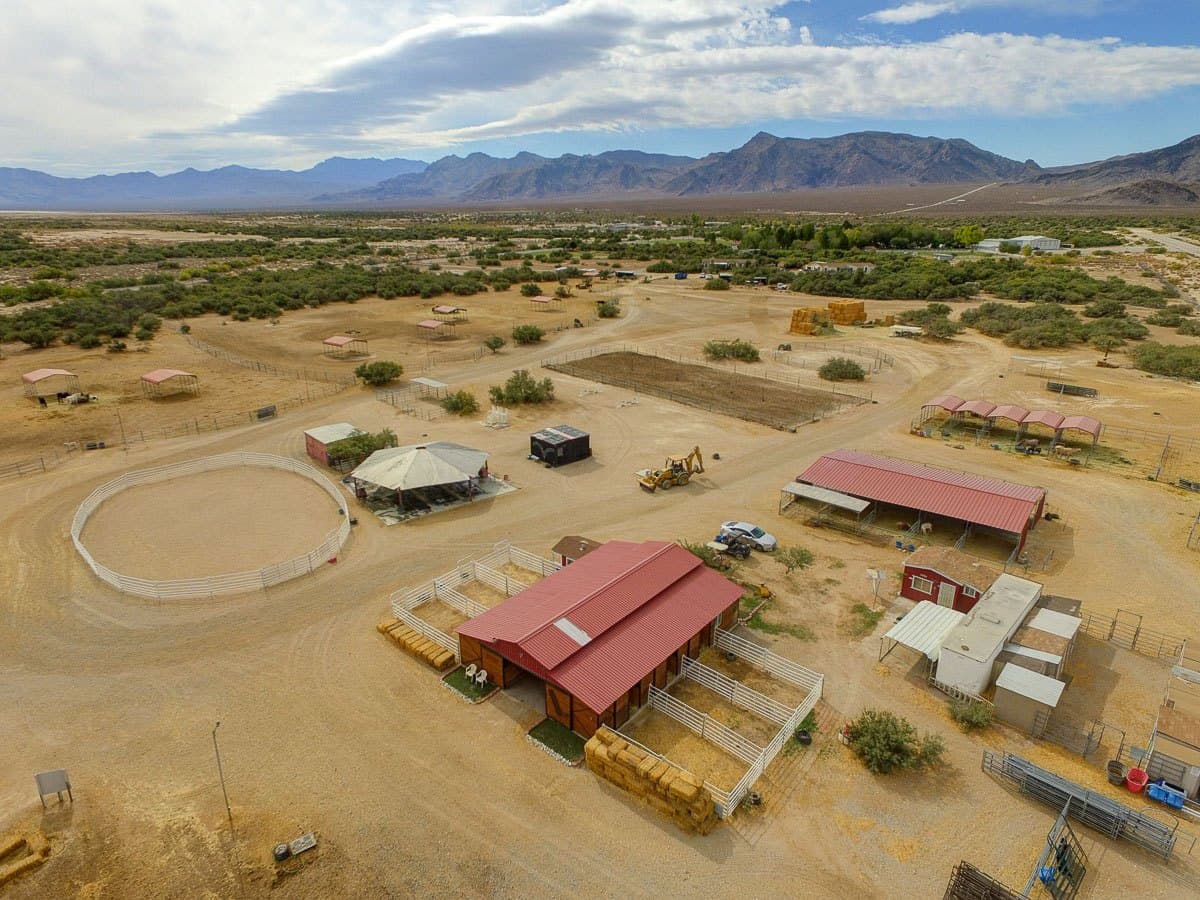 An aerial view of a desert ranch with a circular corral, red-roofed buildings, fences, and distant mountains