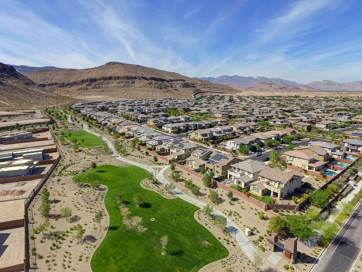 Drone view of a suburban neighborhood with a curved green park, homes, desert landscaping, and mountains