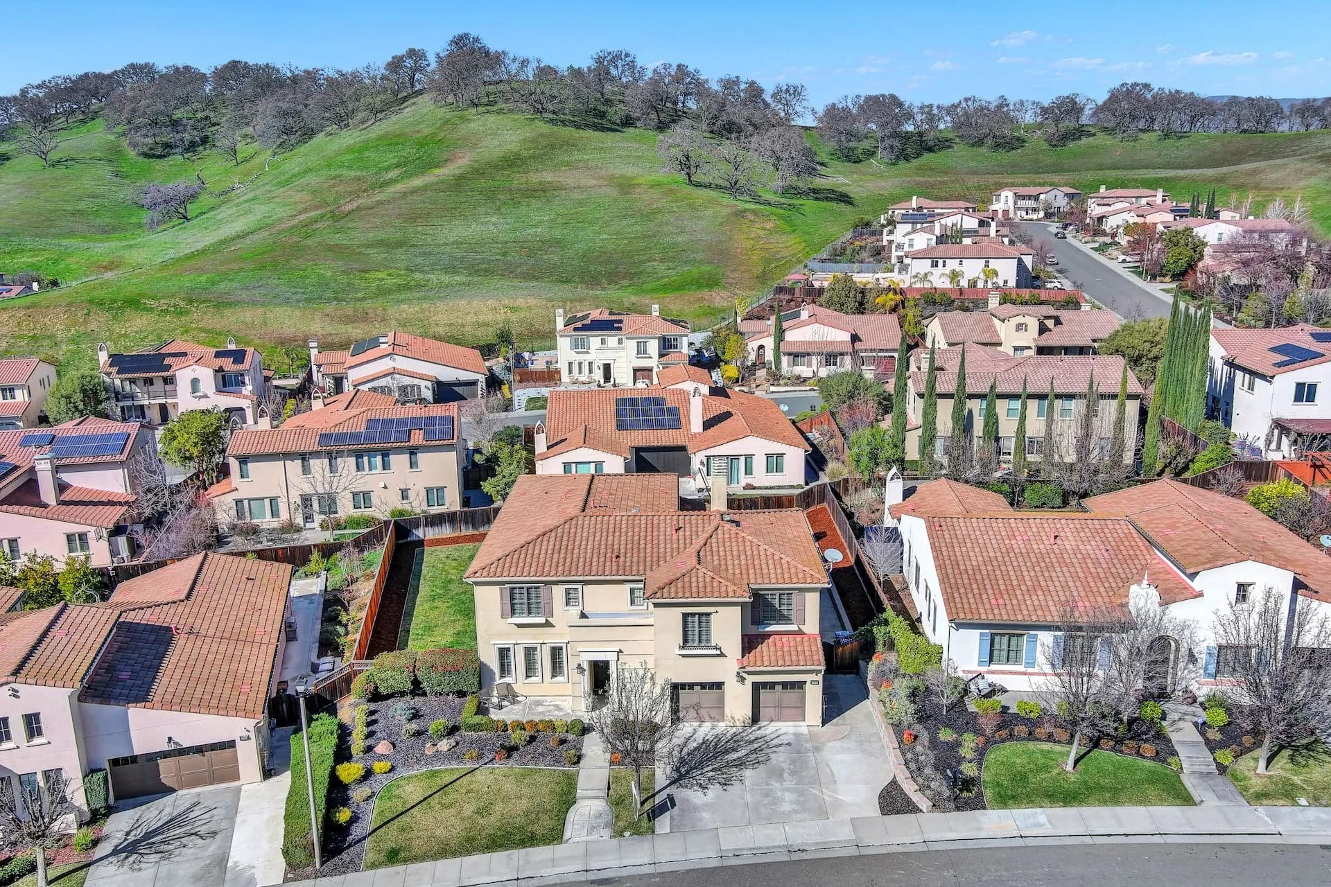 An aerial view of a hillside and nearby neighborhoods with red-tiled roofs, green lawns, and a blue sky